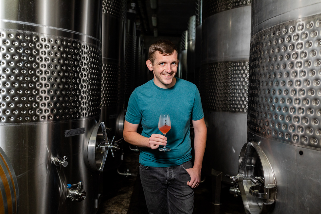 A photo of winemaker Kelby Russell amidst stainless steel tanks, with a glass of red wine in his hand.